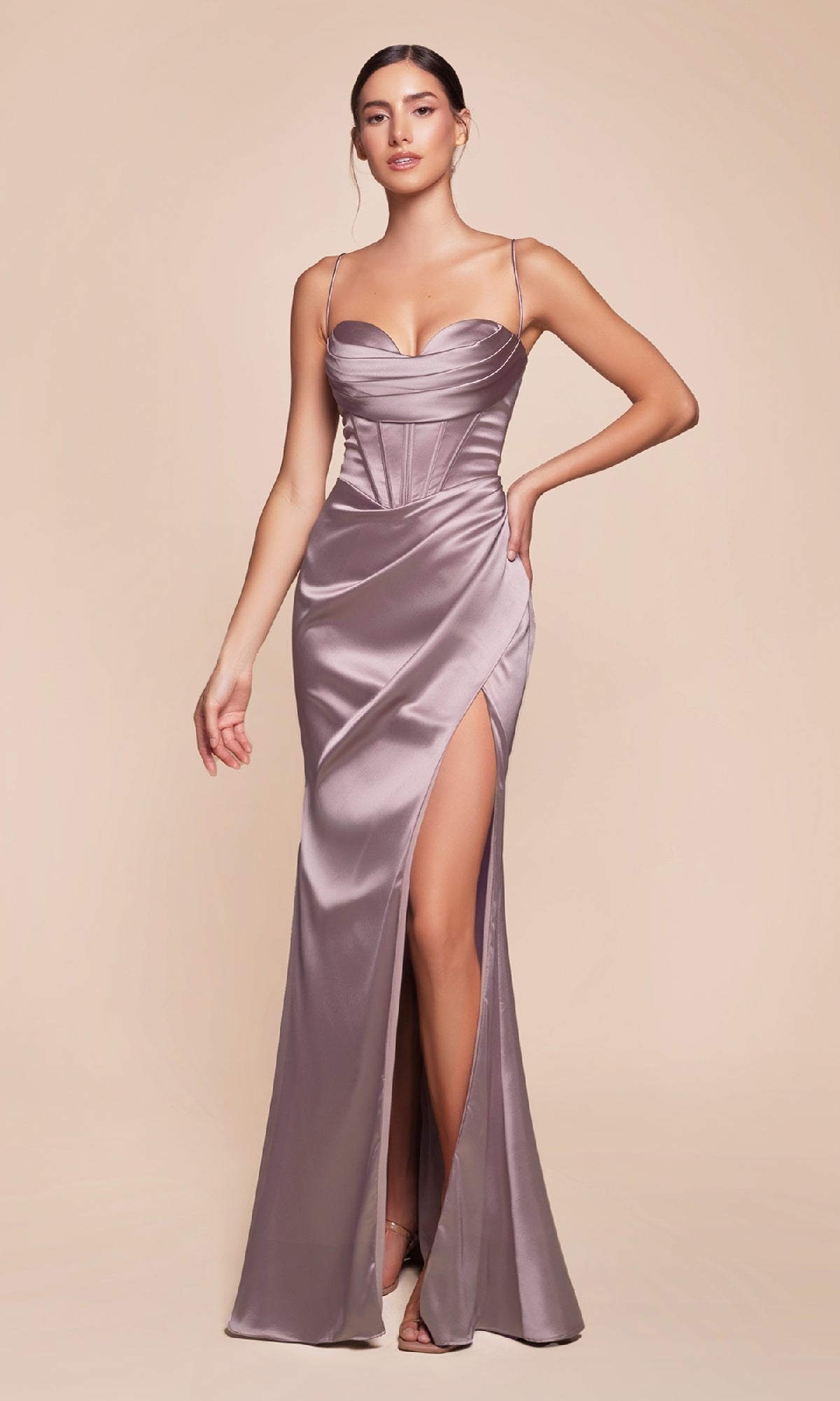 A woman in the Ladivine 7483 Cowl-Neck Backless Corset Prom Dress with thin straps, a fitted bodice, and a high thigh slit poses against a plain beige background.